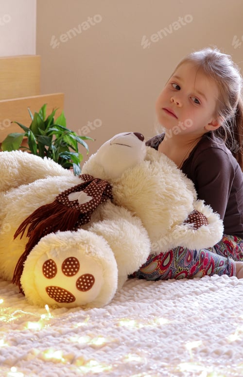 Preview: A little pretty girl looks with a bored look. A teddy bear sits nearby. Bored mood.