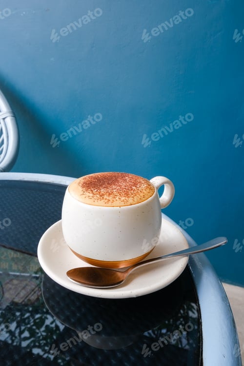 Preview: Cup of fresh cappuccino coffee on glass table with blue bright background.