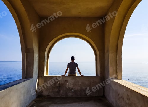 Preview: Silhouette of a man sitting on wall, looking at sea, from behind, inspirational.