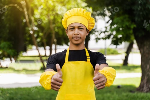 Preview: African teen cook showing thumbs up in yellow apron, perfect bakery promo