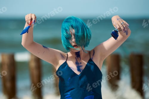 Preview: Artistic blue-haired woman performance artist smeared with blue gouache paints dancing on beach