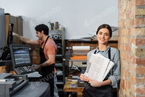 Preview: joyful typographer in apron holding newspapers near colleague on blurred background