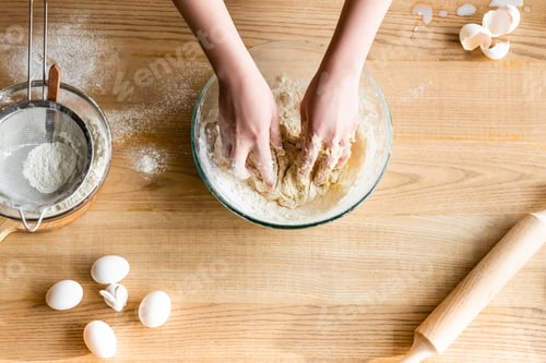 Preview: top view of woman kneading dough near flour in bowl, eggs and figurine with easter bunny