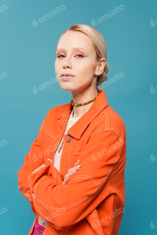 Preview: stylish blonde woman in bright orange jacket posing with crossed arms isolated on blue