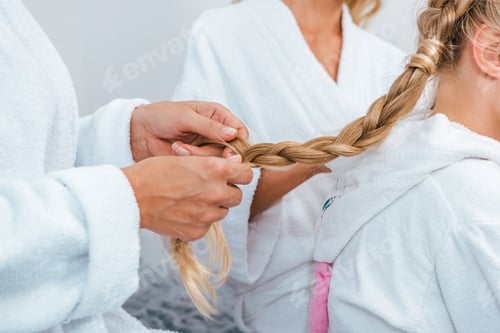Preview: cropped shot of woman braid hair for daughter while they sitting together in bathrobes