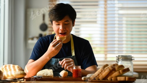 Preview: Attractive young asian man eating a slice of bread with jam for breakfast in kitchen.