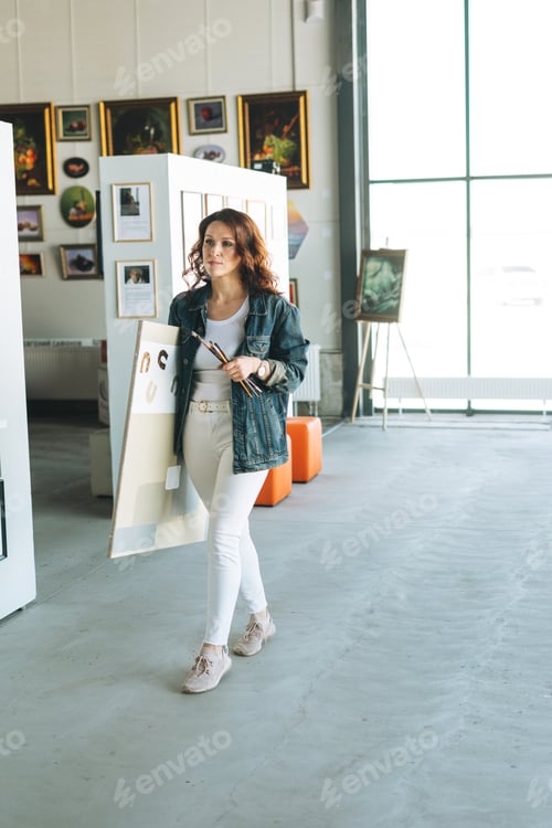 Preview: Charming young woman artist in denim jacket with paint brushes and painting on canvas in art gallery