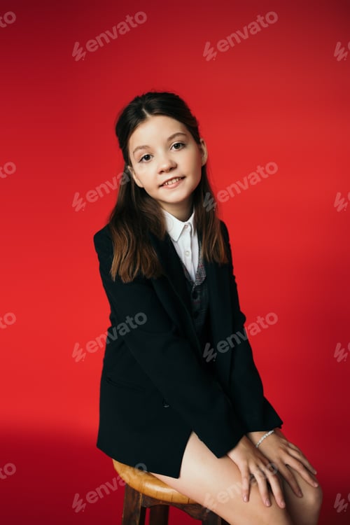 Preview: smiling brunette girl in black jacket sitting on stool and looking at camera on red background