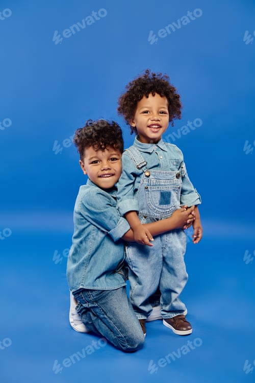 Preview: happy african american preschooler boy in denim clothes embracing toddler brother on blue background