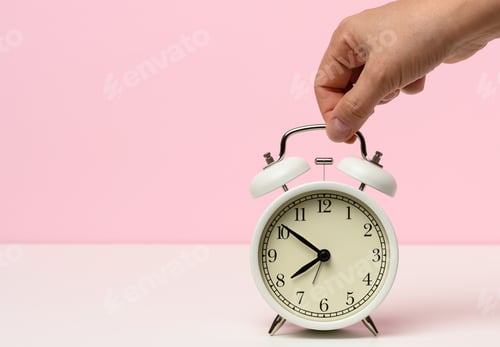 Preview: Female hand holds a white metal alarm clock on a white table, time is ten to eight in the morning