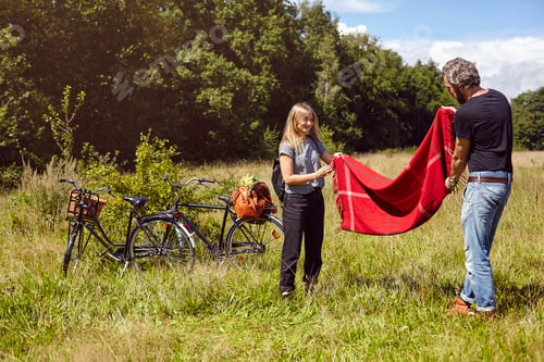 Preview: Two people setting up a picnic blanket next to bikes in a sunny meadow.