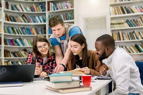 Preview: Four positive smiling multiethnic boys and girls sitting at the table in modern college library and