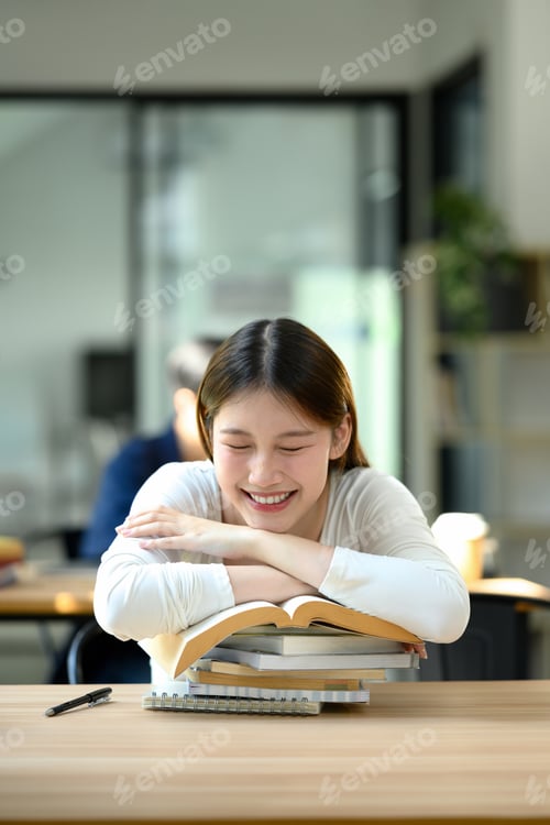 Preview: Smiling young woman taking a break during study time resting her head on a stack of books