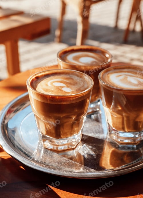 Vista previa: Tres vasos de enfoque selectivo con café con espuma de leche en una bandeja de metal en una cafetería en un día soleado.