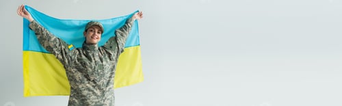 Preview: Happy servicewoman in camouflage uniform holding ukrainian flag isolated on grey, banner