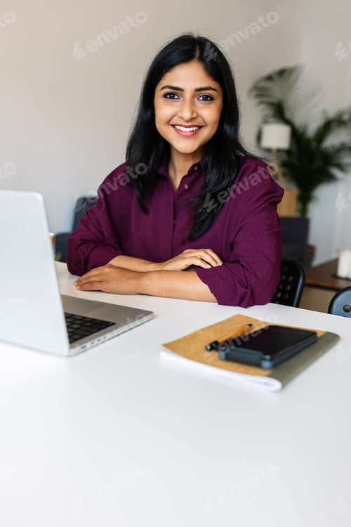 Preview: Vertical shot of young indian woman smile at camera working with laptop at home