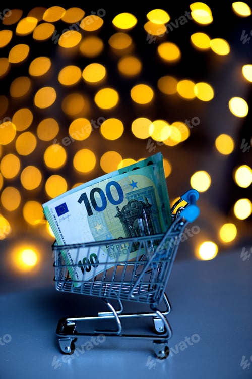 Preview: Christmas shopping online.Trolley with 100 euro notes on the background of blurred bokeh lights.