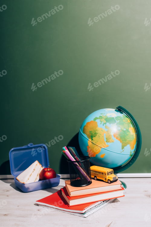 Preview: globe near school bus model and pen holder on books near lunch box on desk near green chalkboard