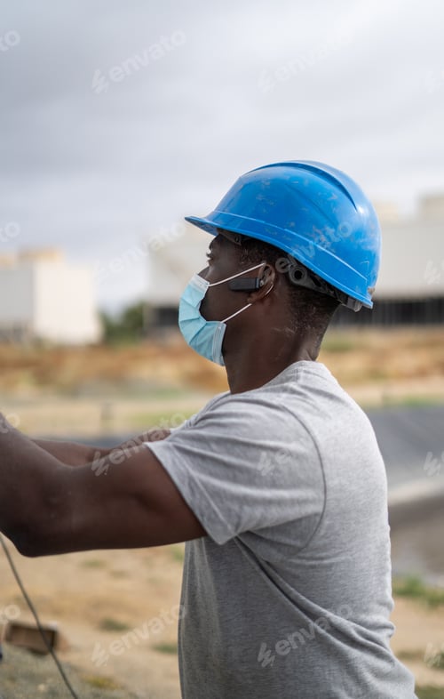 Preview: Afro-American builder wearing a blue helmet and a face mask while working