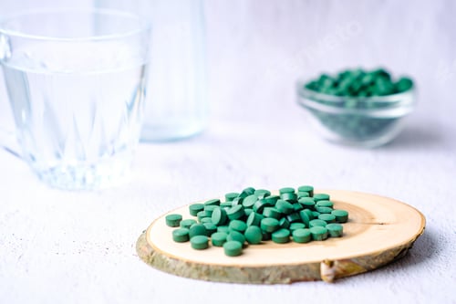 Preview: Green round spirulina pills on natural wood plate on a simple minimalistic gray concrete background.