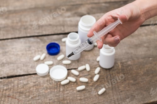 Preview: Hand holds syringe above scattered pills and bottles on wooden surface during medication preparation