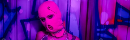 Preview: anonymous woman in pink balaclava looking at camera near colorful graffiti in purple neon light,