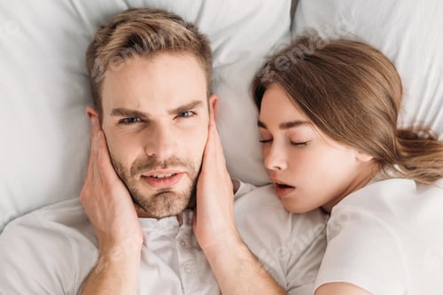 Preview: displeased man plugging ears with hands and looking at camera while lying in bed near snoring wife