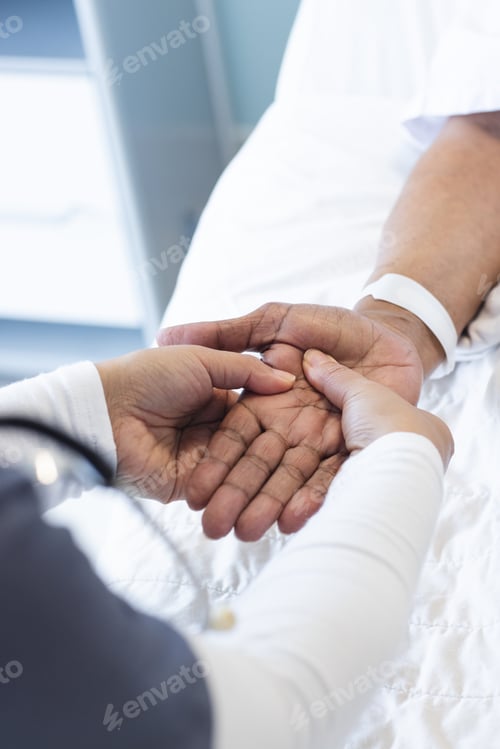 Preview: Hands of diverse female doctor holding hand of senior female patient in hospital room