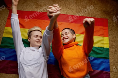 Preview: Happy young lesbian couple lying above lgtb rainbow flag with raised hands up