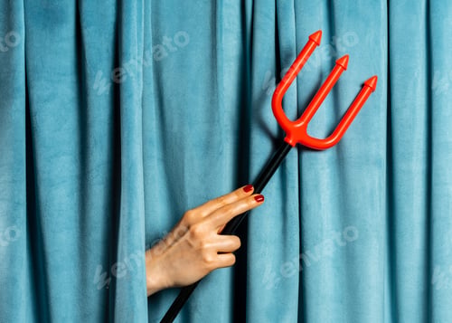 Preview: Female hand hold red devil's trident against blue velvet curtains