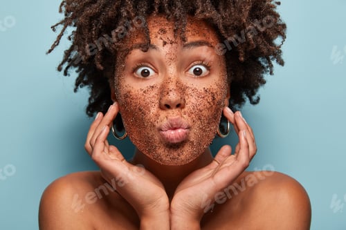 Preview: Close up portrait of African American woman with opened eyes, keeps lips folded, touches cheeks, has