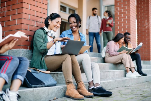 Preview: Happy multi-ethnic female students having fun while using laptop in front of university building.