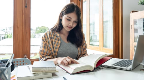 Preview: Young woman student reading textbook and doing homework assignment after using laptop to learning