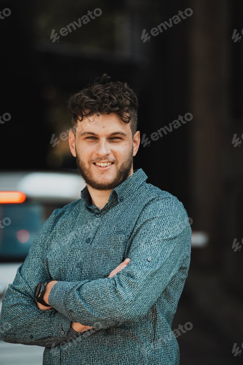 Preview: A successful young businessman in a shirt, with crossed arms, poses outdoors, confident expression