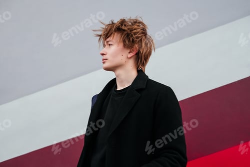 Preview: Thinking young man with birthmarks, moles, wart on face in black coat standing in front of wall