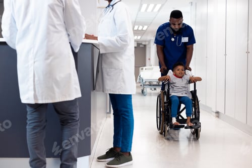 Preview: African american male doctor pushing boy patient in wheelchair in busy hospital corridor