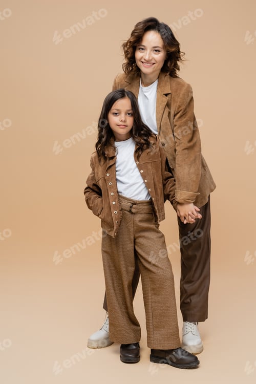 Preview: cheerful woman holding hand with daughter posing in trendy autumn outfit on beige background