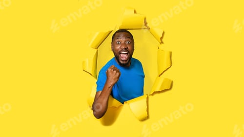 Preview: Excited african american man raising clenched fist and opening mouth, breaking through yellow paper