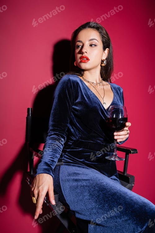 Preview: seductive woman in elegant dress sitting with glass of wine and looking away on red background
