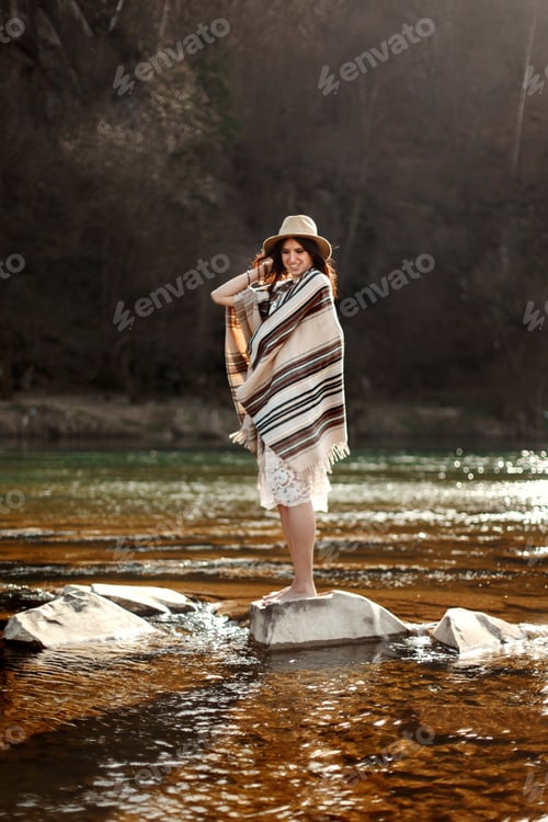 Preview: beautiful woman traveler back standing on rocks in river, wearing hat and poncho