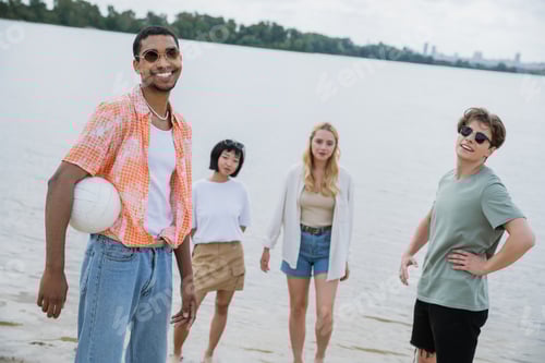 Preview: african american man with ball smiling at camera near multiethnic friends on beach