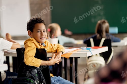 Feliz estudante negro do ensino fundamental sentado na sala de aula e olhando para a câmera.