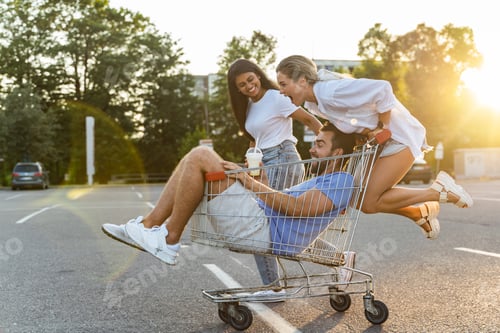 Preview: Three diverse friends having fun and riding shopping cart on parking lot near supermarket