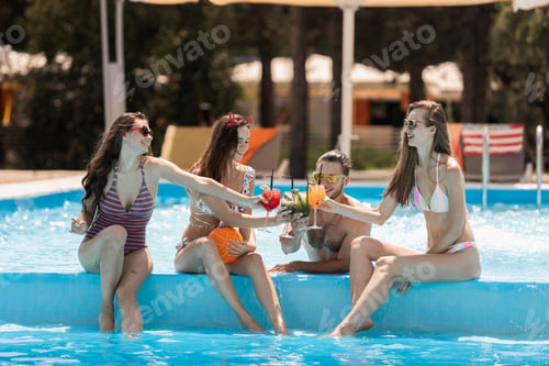 Preview: Young stylish guy and three girls sit on the side and drink cocktails of the swimming pool on the