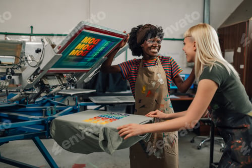 Preview: Smiling interracial print shop female workers silkscreen printing t-shirt.