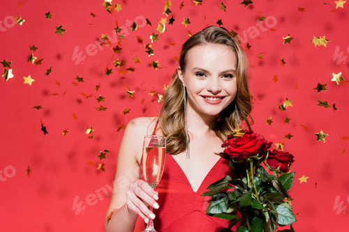 Preview: happy woman holding roses and glass of champagne near falling confetti on red