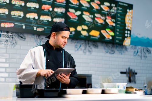 Preview: Thoughtful Sushi Chef with a tablet in his hands near the kitchen table on which products for rolls