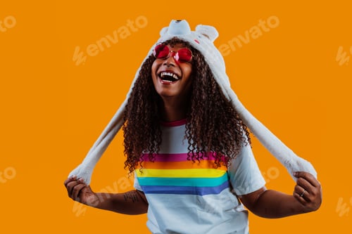 Preview: Young woman wearing funny hat and rainbow t-shirt is laughing on orange background
