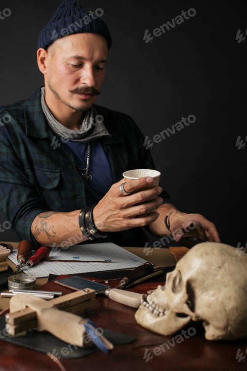 Preview: Leather Craftsman using smart phone and holding cup of coffee indoors