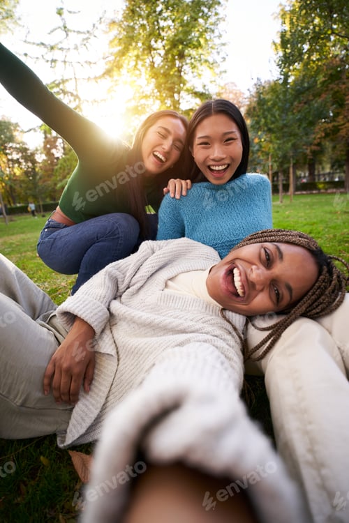 Preview: Vertical photo of Three multiracial girls lying on the grass taking selfie looking at camera.